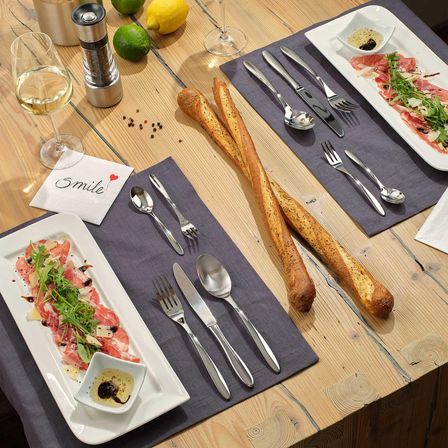 Ein Tisch mit einem Glas Wein, einem Teller mit Fleisch und Grünzeug, Brot und Besteck gedeckt.