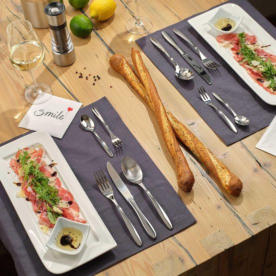 Ein Tisch mit einem Glas Wein, einem Teller mit Fleisch und Grünzeug, Brot und Besteck gedeckt.