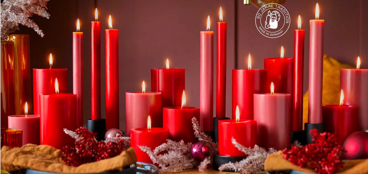 A collection of red candles are displayed on a table, lit with a warm glow. A red ornament is placed on the table, and a logo is displayed on the wall.