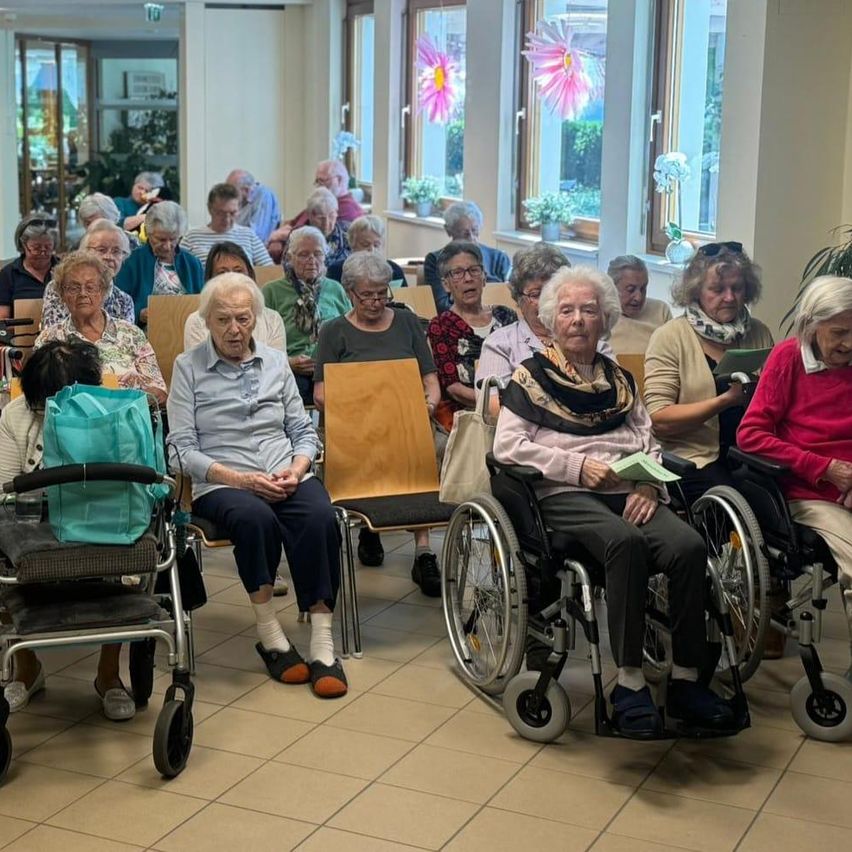Ein Raum voller älterer Menschen, die auf Stühlen und Rollstühlen sitzen. Einige halten Papiere. Blumen sind auf den Fensterbänken.