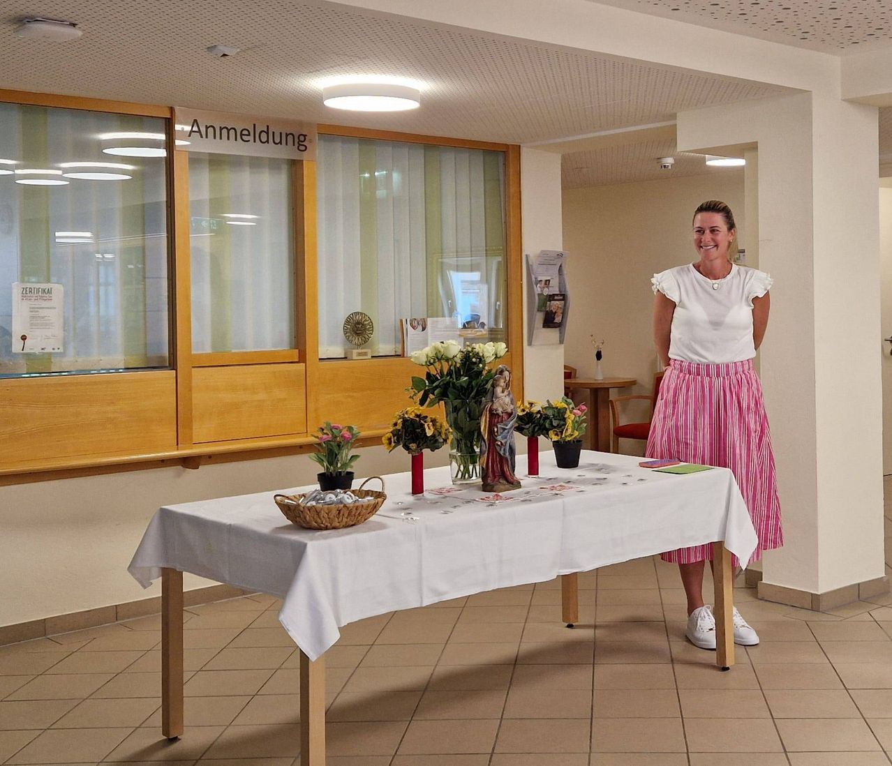 Eine Frau steht neben einem Tisch mit Blumen und Kerzen. Der Tisch ist mit einem weißen Tuch bedeckt. Hinter ihr befindet sich eine Glastür mit einem Schild, das 'Anmeldung' besagt.