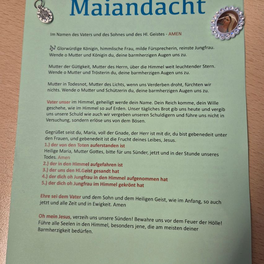 Ein grünes Papier zeigt ein religiöses Gebet mit dem Titel 'Maiandaht'. Es enthält einen silbernen Anhänger und einen Bilderrahmen mit einer Frau. Das Gebet richtet sich an Maria und preisen sie als glorreiche Königin, himmlische Frau, milde Fürsprecherin und reine Jungfrau. Es bittet um ihren mitfühlenden Blick, der sich ihnen zuwendet.