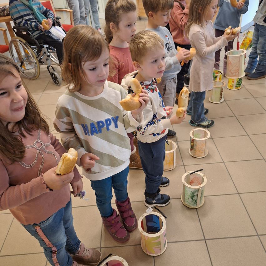 Mehrere Kinder stehen in einer Reihe und halten Brot in den Händen. Sie tragen Winterkleidung und Stiefel. Der Raum hat einen gefliesten Boden und im Hintergrund ist ein Rollstuhl zu sehen.