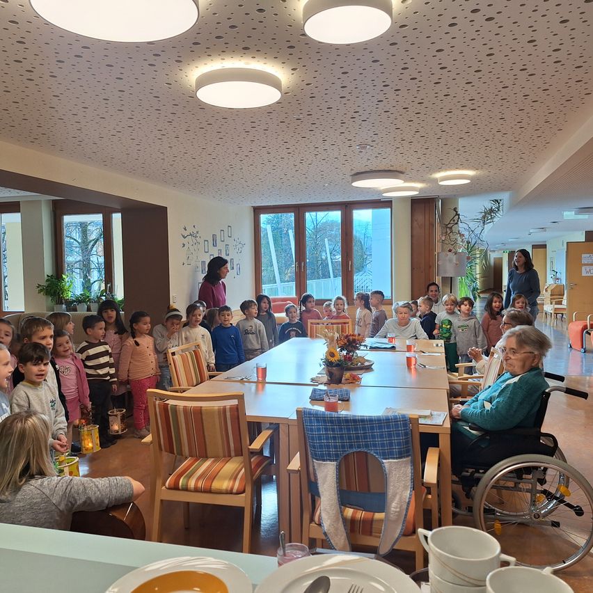Eine Gruppe von Kindern steht in einem Raum, wahrscheinlich einem Klassenzimmer, mit einer älteren Frau im Rollstuhl. Sie scheinen zu singen oder zu sprechen. Der Raum hat große Fenster und einen Tisch mit Blumen und Tassen.