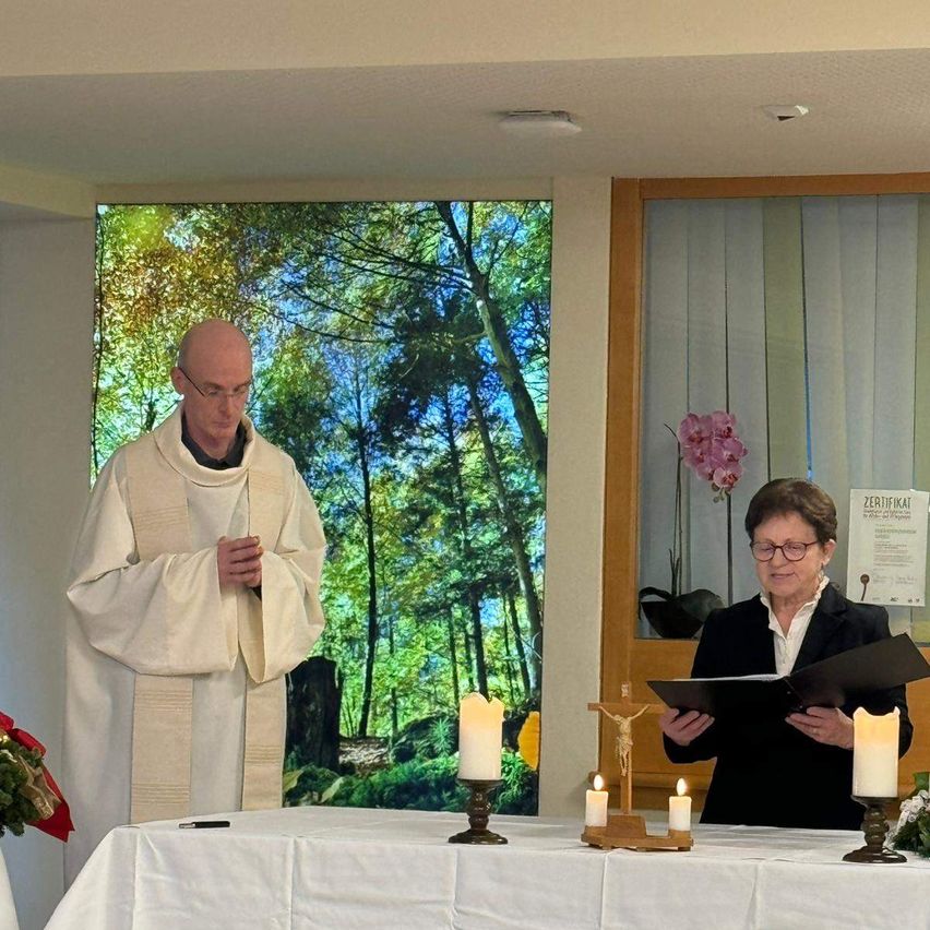 A priest stands praying in a church with candles and flowers, while a woman reads from a book.