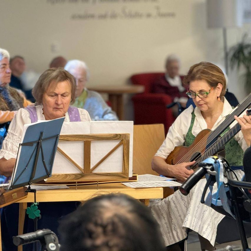 Two women play instruments, one a guitar, the other a lute, while reading sheet music. They perform for an audience in a room.