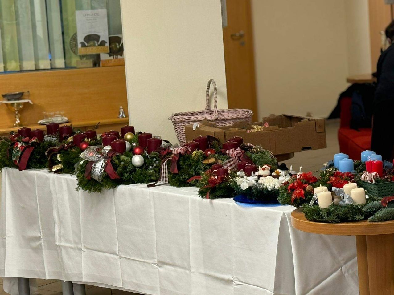 A white table covered with a white tablecloth, decorated with Christmas wreaths, candles, and ornaments. A basket and a box are placed behind the table. A wooden bench is on the left.