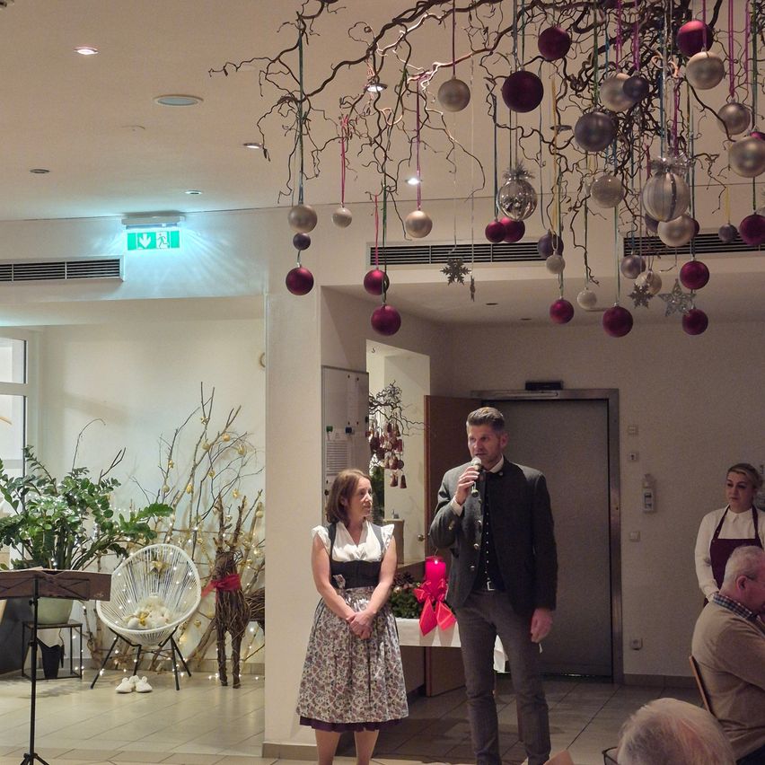 A man and a woman stand before a crowd in a decorated room, possibly for a ceremony. They are near a table with a red ribbon. A Christmas tree with ornaments hangs from the ceiling.