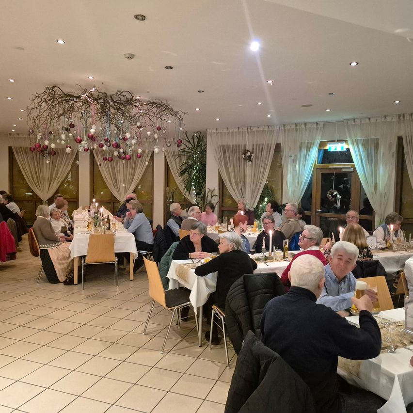 A dining room with tiled flooring, long tables covered in white tablecloths. People are sitting around the tables, some are having conversations. A chandelier with Christmas decorations is hanging from the ceiling.
