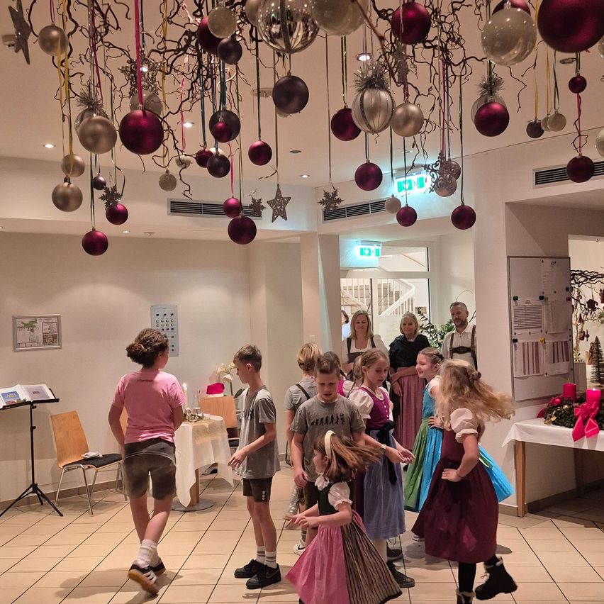 A group of children dance in a room decorated with Christmas ornaments and adults observe. Tables with flowers and books are present.