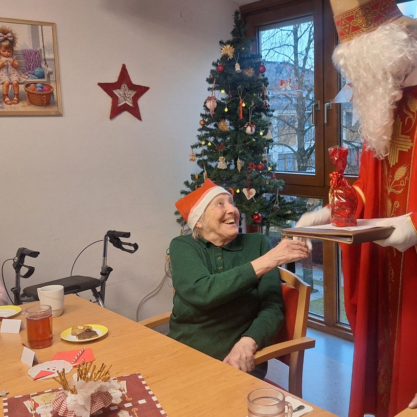 An elderly woman in a green sweater and Santa hat sits at a table, smiling as she receives a gift from a person dressed as Santa Claus, with a Christmas tree and a picture frame on the wall behind them.