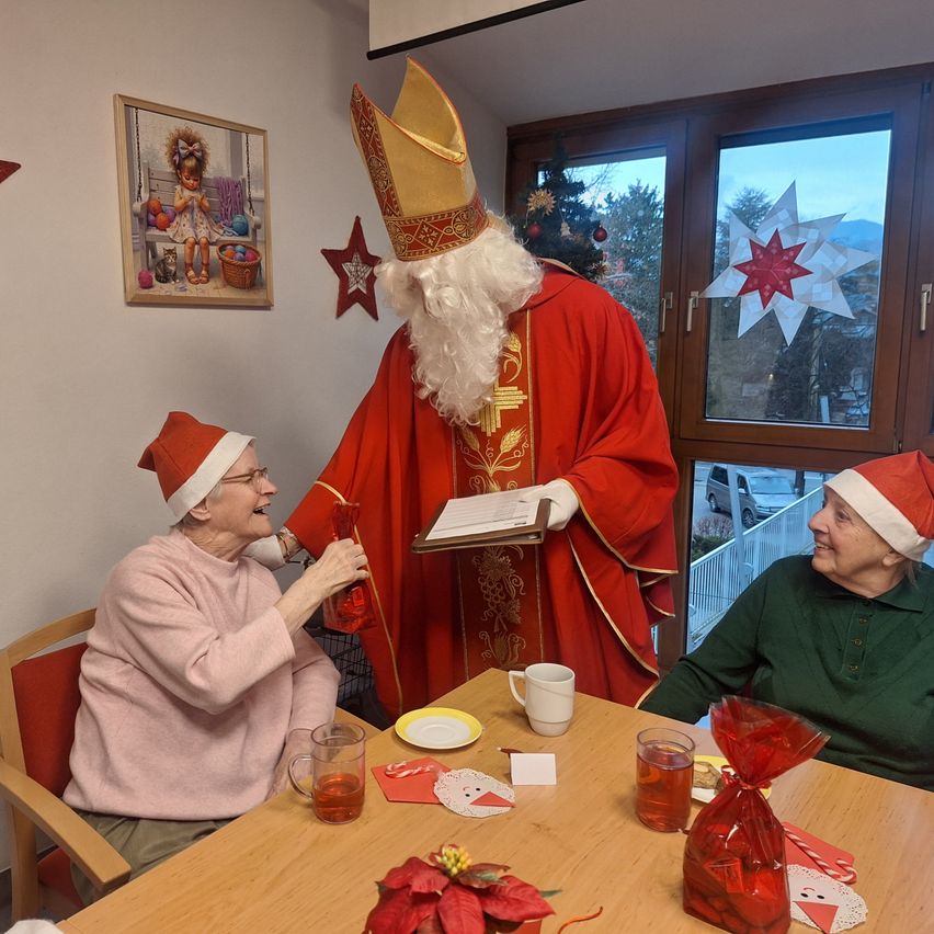 Two elderly women sit at a table with drinks and a man dressed as St. Nicholas holding a book. Behind them, a window displays a Christmas decoration.