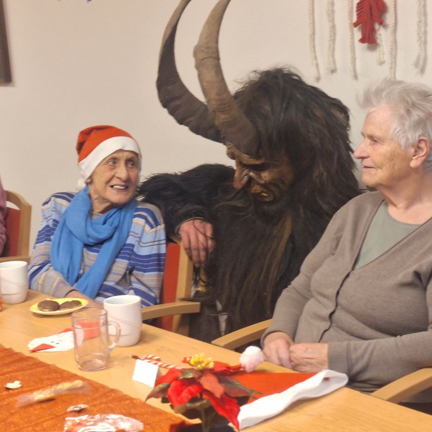 Two elderly women sit at a table with a man in a demon costume, smiling. The table has cups, a plate with cookies, and a red flower. A white wall with decorations is behind them.