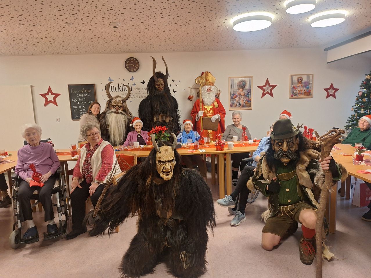 A group of elderly individuals in a room are dressed in various costumes for a Christmas party. A person in a bear costume is standing in the center.