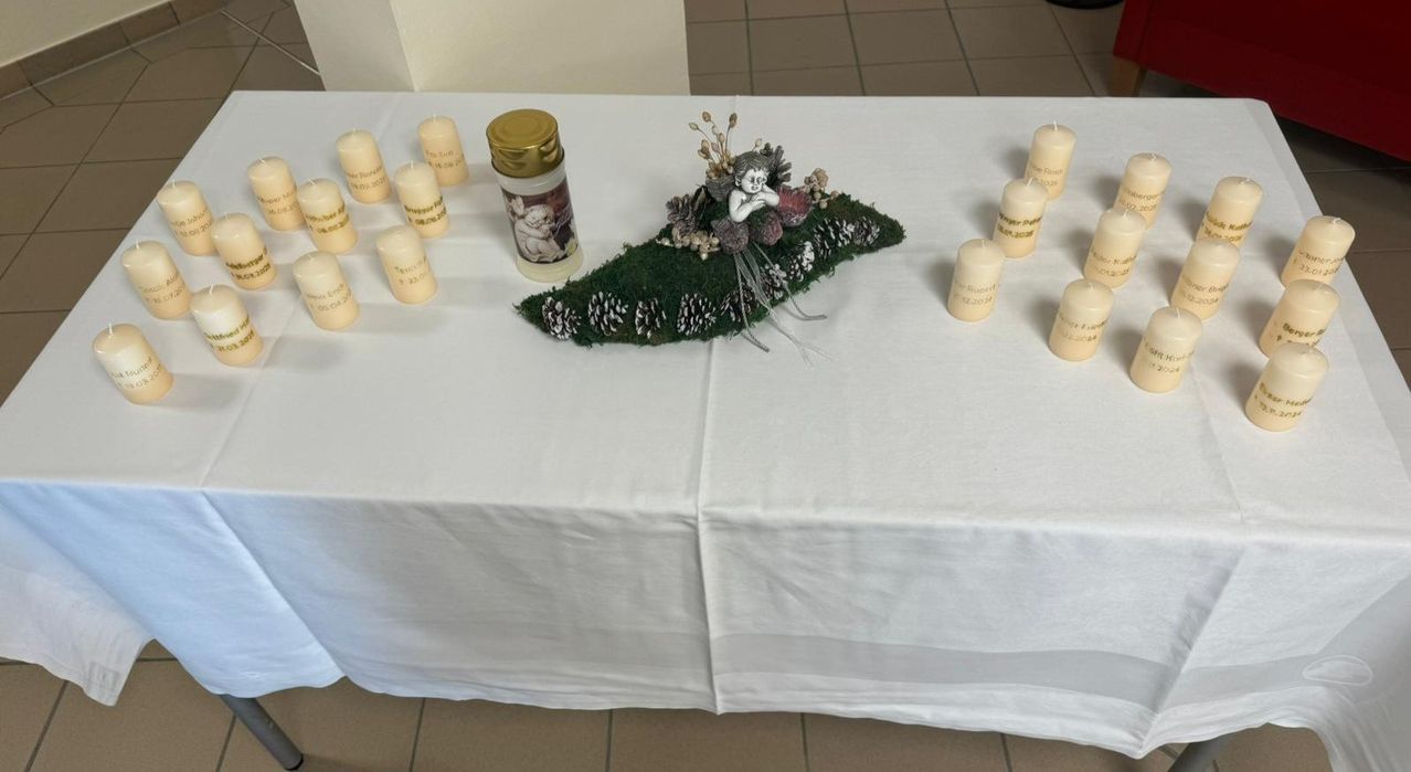 A white table covered with a white cloth has several white candles and a decorative item. The candles have names and dates on them. The decorative item is a green and brown design with a statue on top.