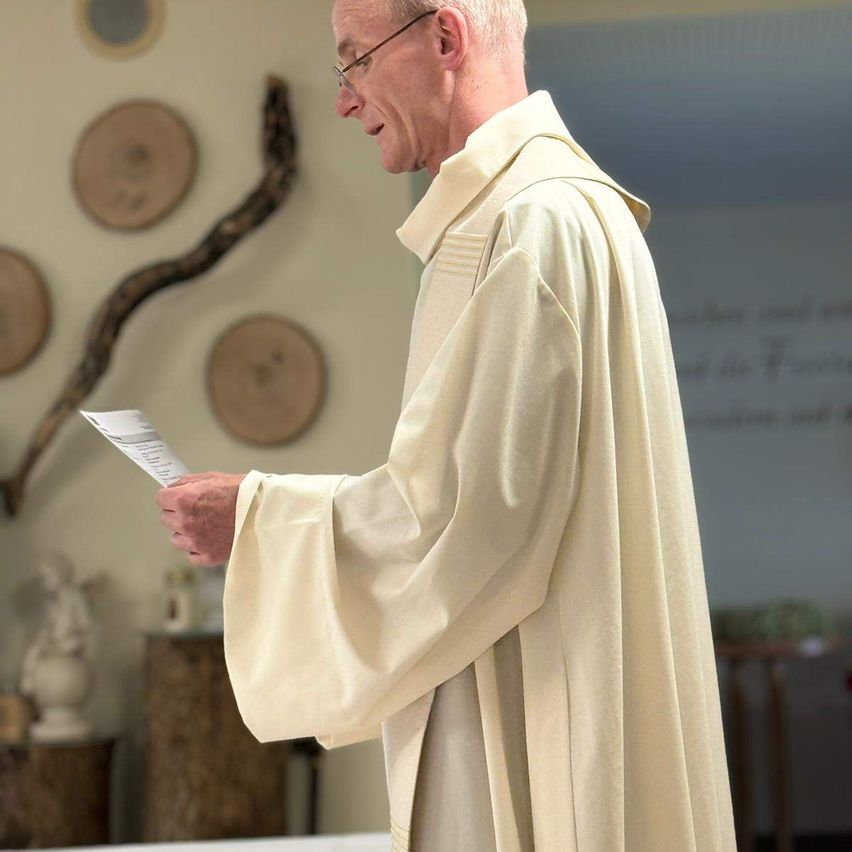 A priest stands in a room, holding a paper and reading, with a white robe and glasses, surrounded by wooden decorations on the wall.