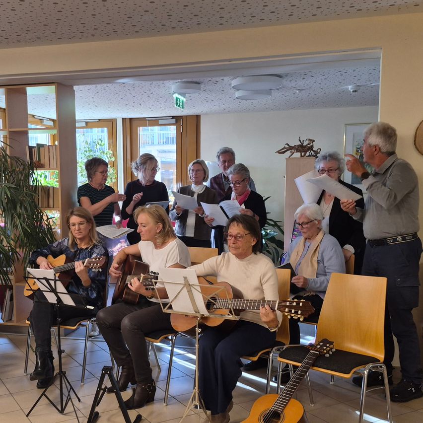 A group of adults is practicing music with guitars and sheet music in a room with bright lighting and large windows.