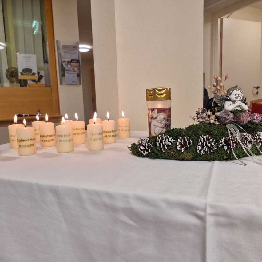 A table covered with a white cloth holds multiple lit candles and a floral arrangement with a cherub figure. Behind the table is a white wall with a framed picture.