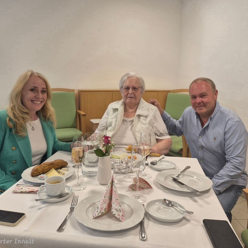 Drei Personen sitzen um einen Tisch mit Weingläsern und Tellern. Eine Frau trägt einen grünen Pullover und eine Halskette, während ein Mann ein blaues Hemd trägt. Der Tisch ist mit Brot, Käse und Kaffee gedeckt. Hinter ihnen stehen grüne Stühle.