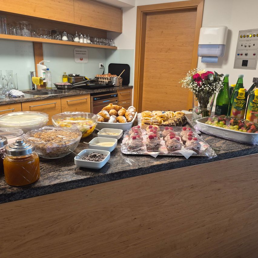 Ein Buffettisch mit verschiedenen Gebäckstücken, Früchten und Getränken. Der Tisch ist mit Desserts, Früchten und Getränken gefüllt, mit einer Blumenarrangement in der Mitte.
