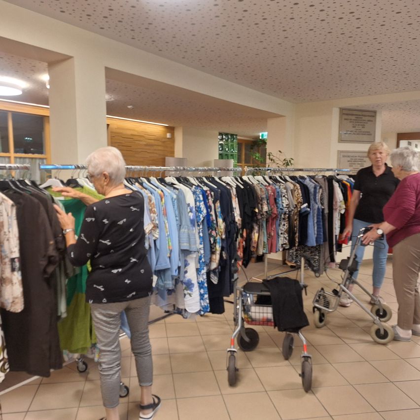 Three elderly women are browsing through a clothing store with a rack of clothes in front of them. The woman in the middle is holding a shirt and looking at it. The woman on the right is holding a walker. The woman on the left is looking at the clothes on the rack.