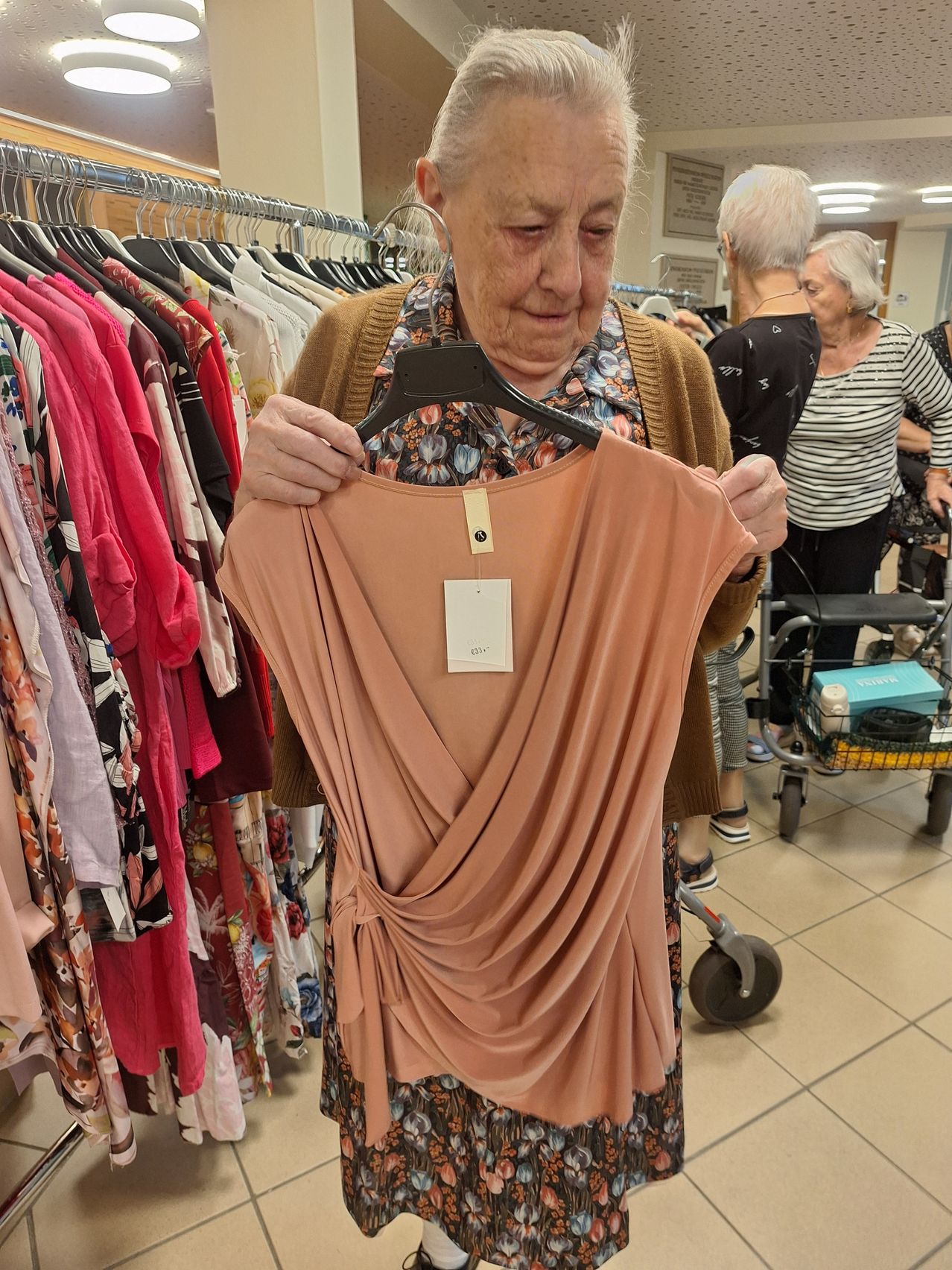 An elderly woman examines a peach-colored blouse in a clothing store, with racks of colorful clothes in the background.