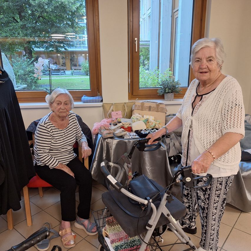 Two elderly women are in a room with a table full of items, one holding a walker. The woman on the left is sitting on a chair, and the other is standing next to a table with various objects.
