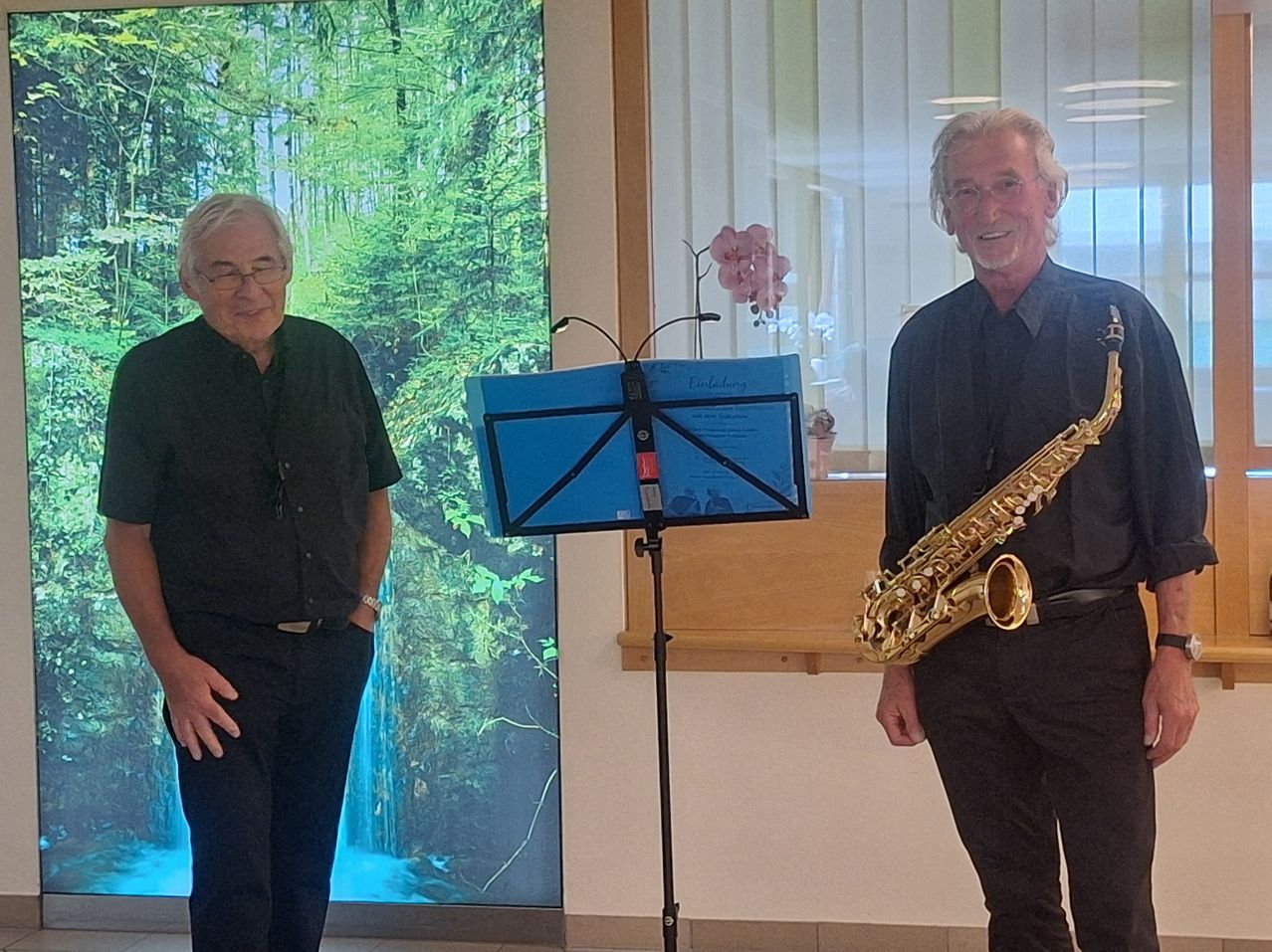 Two older men stand next to each other, one holding a saxophone, the other with a music stand. They are in front of a large window with a forest background.
