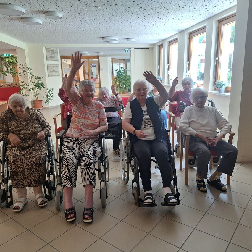 A group of elderly women in wheelchairs are smiling and raising their hands in a room with white tiles and windows.