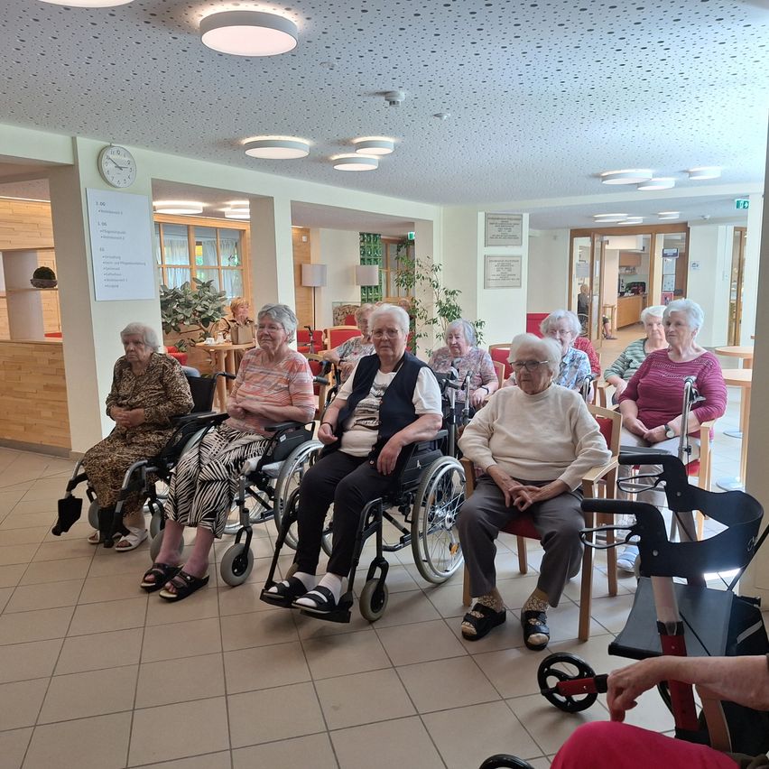 A group of elderly women in wheelchairs and chairs, sitting in a room with tiled floors and ceiling lights. A clock and plants are visible in the background.