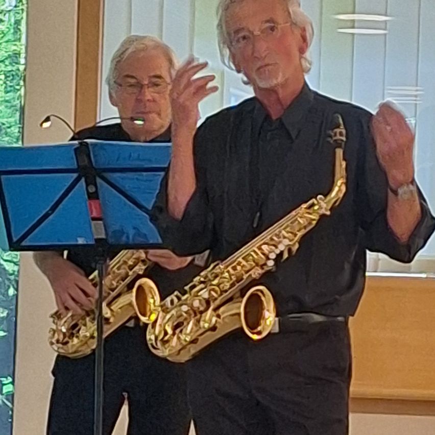 Two men are playing saxophones, one holding a microphone and the other gesturing while playing. They are standing in front of a music stand.
