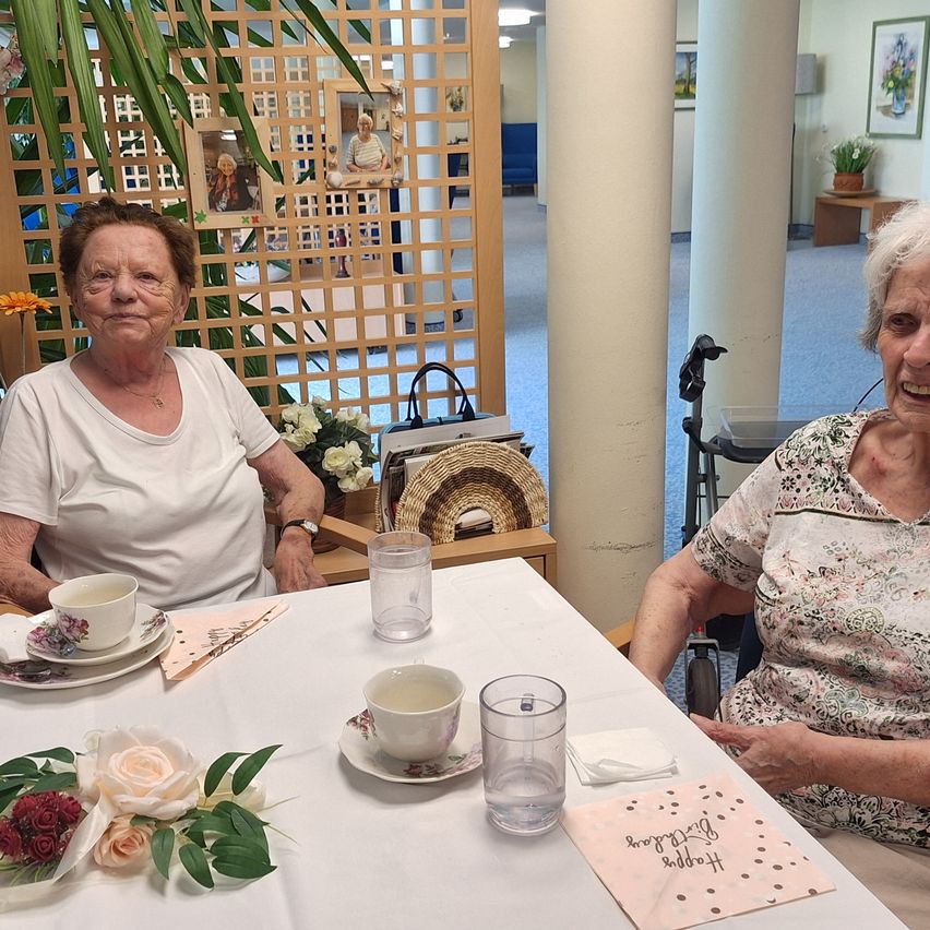 Zwei ältere Frauen sitzen an einem Tisch mit Blumen, Tassen und Untertassen. Eine Frau lächelt, während die andere in die Kamera schaut. Dahinter befindet sich eine Holzabtrennung mit Bildern und einer Pflanze.