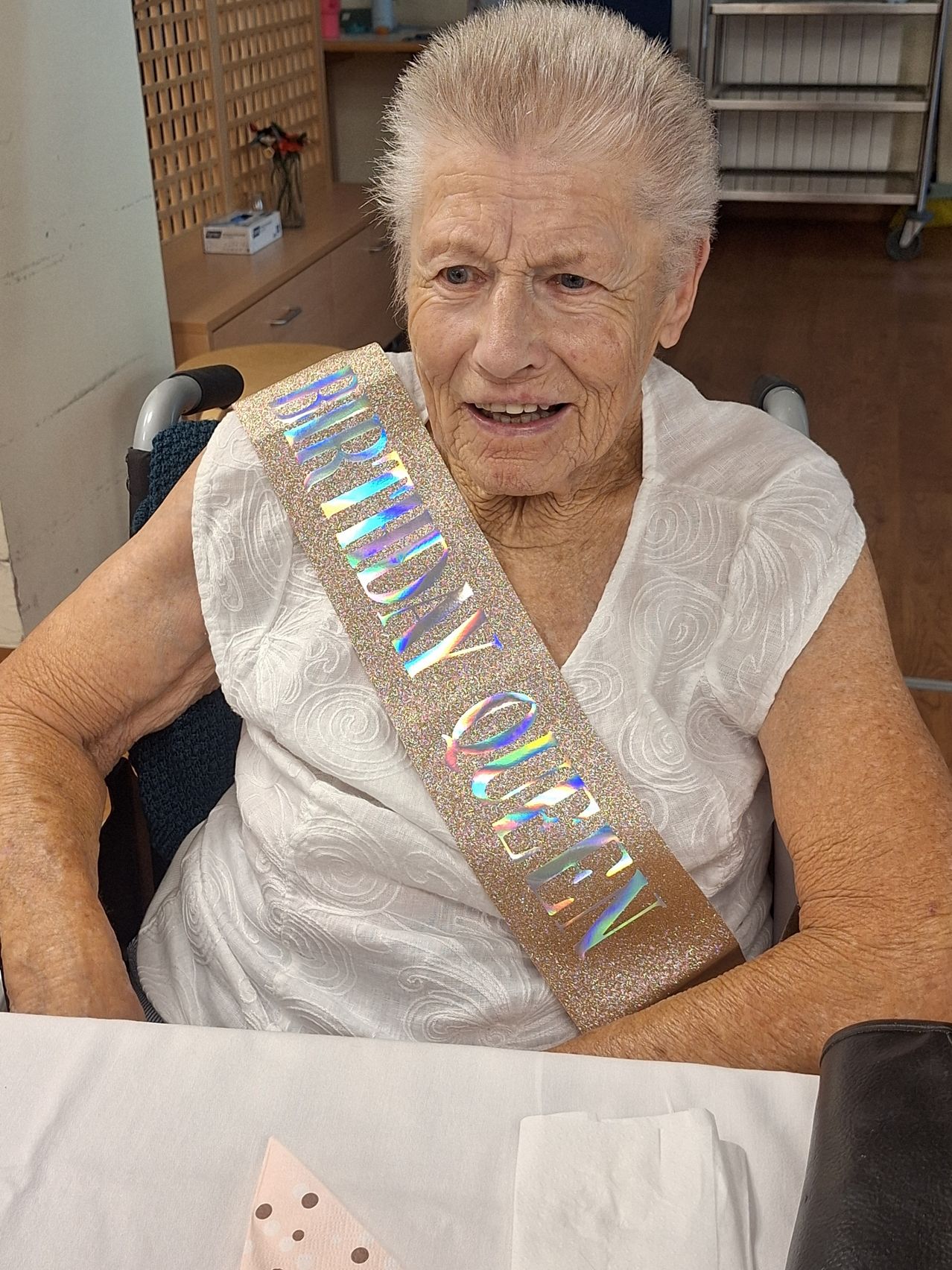 Eine ältere Frau, die ein weißes Oberteil und eine Geburtstagsschleife mit der Aufschrift 'GEBURTSTAGS-QUEEN' trägt, sitzt lächelnd in einem Stuhl.