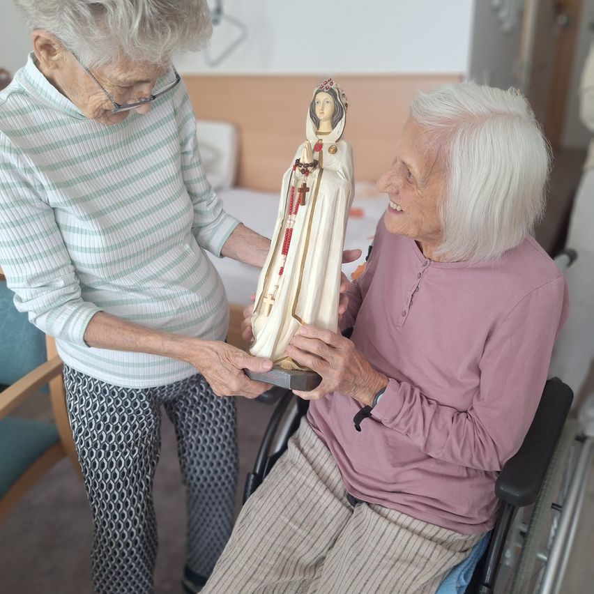 Zwei ältere Frauen betrachten eine Statue einer Heiligen. Die Frau im Rollstuhl lächelt und hält die Statue mit beiden Händen. Die andere Frau steht neben ihr, trägt eine Brille und hält die Basis der Statue.
