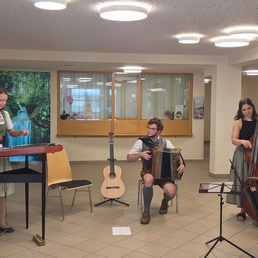 Drei Musiker spielen in einem Raum mit gefliesten Böden. Ein Mann spielt Akkordeon, eine Frau spielt Cello und eine andere Frau spielt Klavier. Der Raum hat große Fenster und eine Wandmalerei.