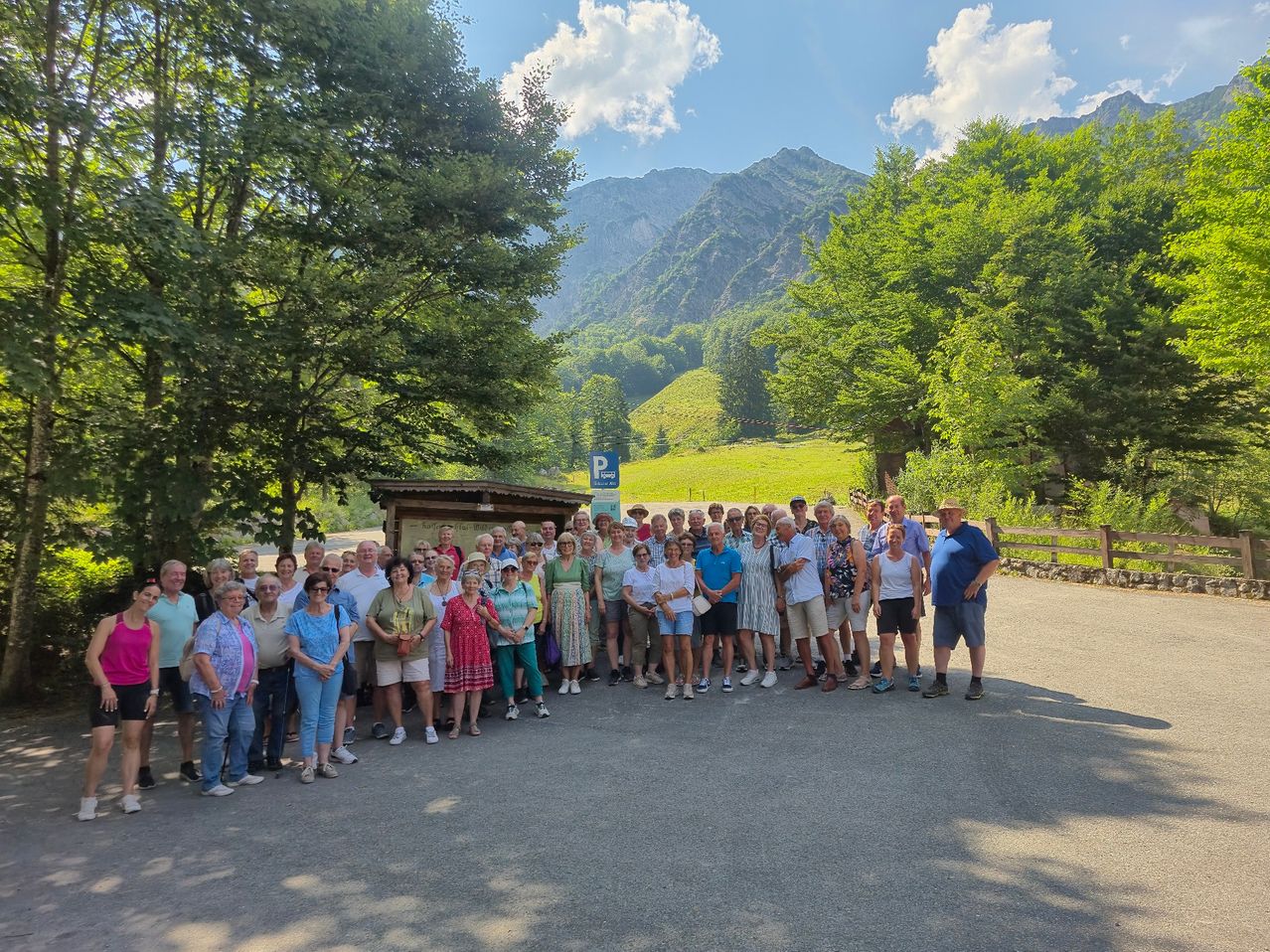 A group of elderly people poses for a photo in a mountain valley, near a parking sign. Trees and a fence are in the background.