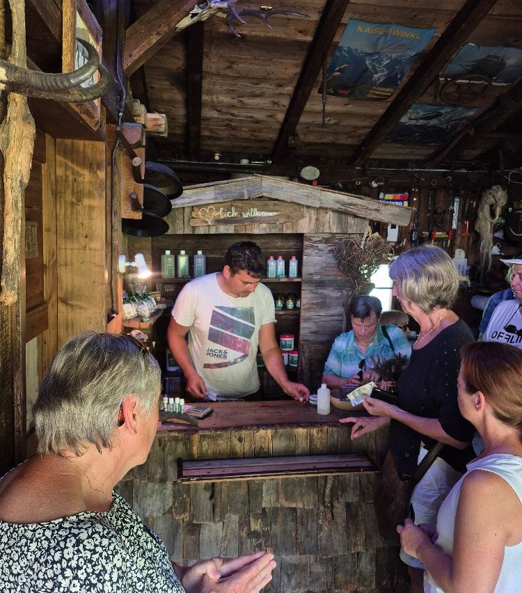 A man in a white t-shirt stands behind a wooden counter in a rustic shop, serving a group of older women. Various bottles and items are on display behind him.