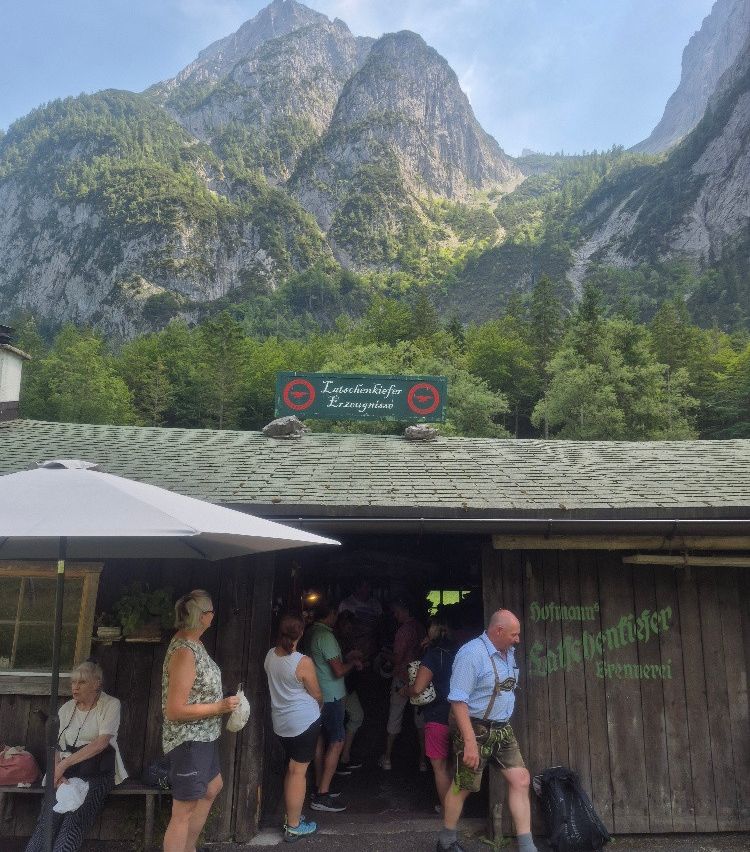 A group of people are gathered outside a rustic wooden building with mountains in the background. A sign reading 'Lagenchliefer' is visible above the entrance.