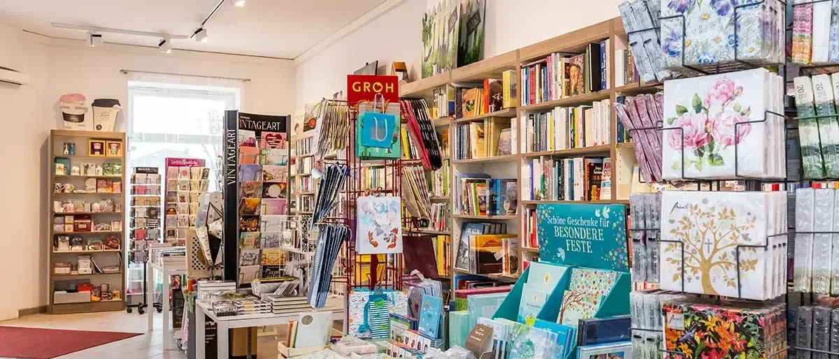 Ein Geschäft voller Bücher, Karten und verschiedener Artikel. Regale sind mit Büchern bestückt, und es sind farbenfrohe Karten ausgestellt. Ein rotes Schild mit der Aufschrift 'Groh'. Ein Schreibtisch steht in der Ecke mit Büchern darauf.