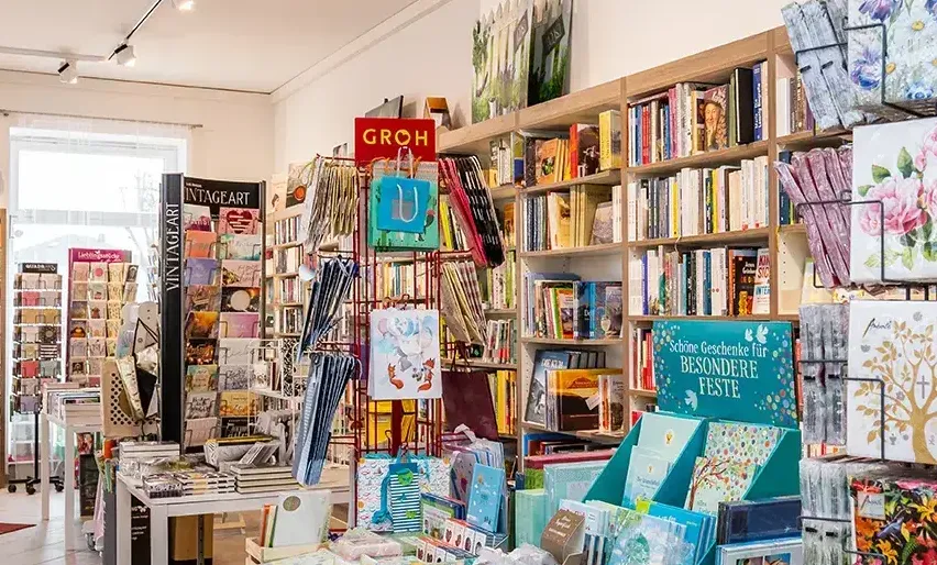 Ein Geschäft voller Bücher, Karten und verschiedener Artikel. Regale sind mit Büchern bestückt, und es sind farbenfrohe Karten ausgestellt. Ein rotes Schild mit der Aufschrift 'Groh'. Ein Schreibtisch steht in der Ecke mit Büchern darauf.