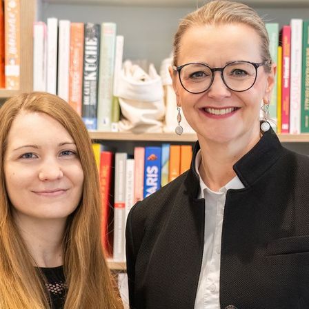 Zwei Frauen lächeln und stehen vor einem Bücherregal. Die Frau links hat lange Haare und die Frau rechts trägt eine Brille und einen schwarzen Mantel.