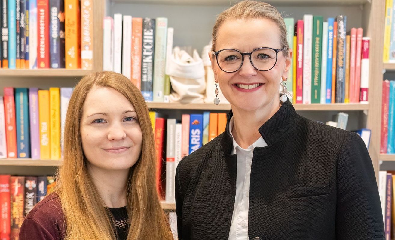 Zwei Frauen lächeln und stehen vor einem Bücherregal. Die Frau links hat lange Haare und die Frau rechts trägt eine Brille und einen schwarzen Mantel.