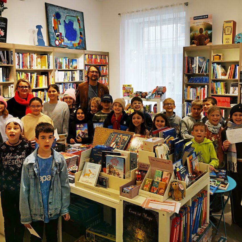 Gruppe von Kindern und Erwachsenen, die hinter einem Büchertisch in einer Buchhandlung stehen, mit Bücherregalen im Hintergrund.
