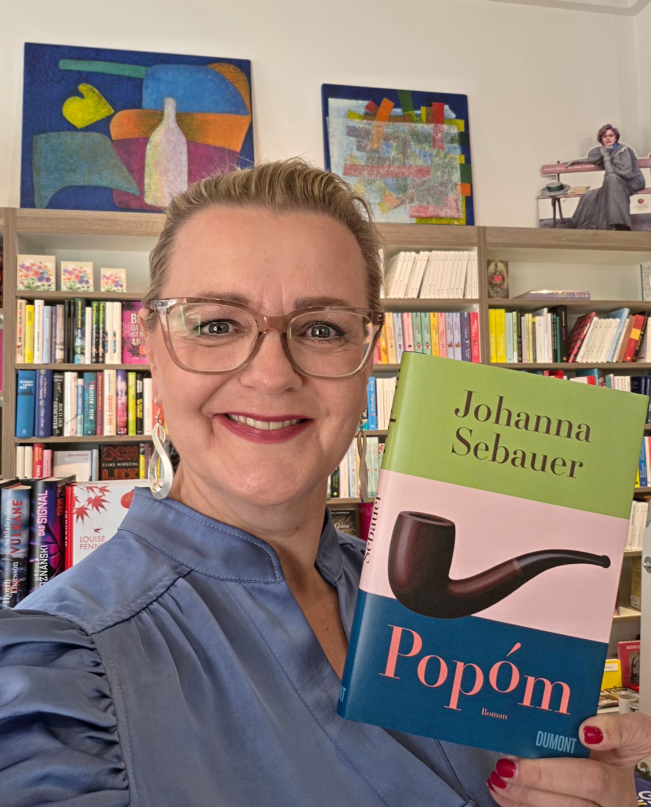 Eine Frau mit Brille hält ein Buch mit dem Titel 'Popom' von Johanna Sebauer hoch. Sie lächelt und steht vor einem Bücherregal.