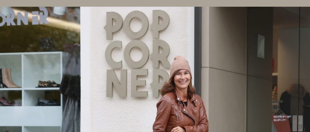 Eine Frau in einer braunen Lederjacke, schwarzem Rock und schwarzen Stiefeln geht vor einem Geschäft mit dem Schild POP CORNER.