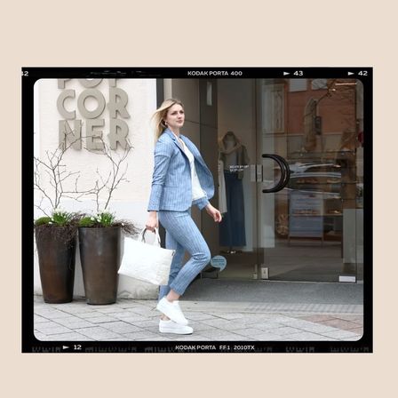 Eine Frau in einem blauen Anzug geht an einem Geschäft namens 'Corner' vorbei. Sie trägt eine weiße Handtasche. Das Schaufenster des Ladens zeigt Dummies und ein Schild mit dem Wort 'Corner'.