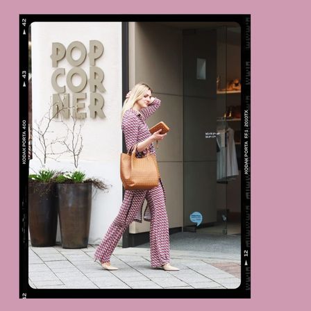 Eine Frau geht mit einer großen Handtasche den Bürgersteig entlang, in der Nähe eines Ladens mit rosa Hintergrund und dem Wort Pop Corner.