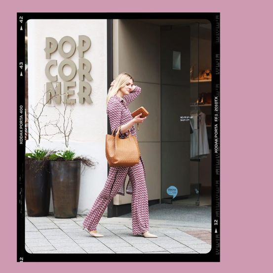 Eine Frau geht mit einer großen Handtasche den Bürgersteig entlang, in der Nähe eines Ladens mit rosa Hintergrund und dem Wort Pop Corner.