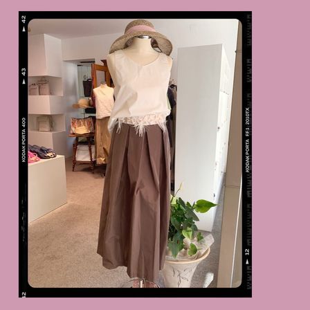 Eine Puppe in einem weißen, ärmellosen Oberteil und einem braunen Rock steht vor einem Spiegel in einem Bekleidungsgeschäft.