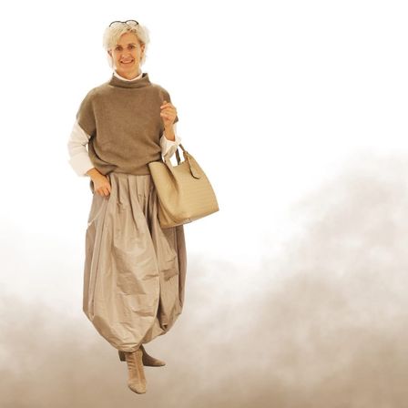 Eine Frau mit weißen Haaren steht in einer wolkenreichen Umgebung, trägt ein beigefarbenes Rollkragenshirt, einen Rock und Stiefel und hält eine beige Handtasche.